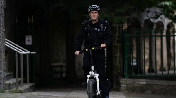 Beyond Paradise Series Two - PC Kelby Hartford (Dylan Llewellyn) in police uniform riding an e-scooter