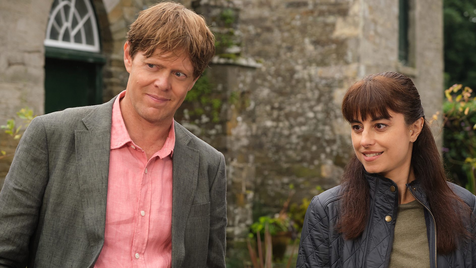 Beyond Paradise Series 2 - Humphrey Goodman (Kris Marshall) and DS Esther Williams (Zahra Ahmadi) stand outside and look at someone off-camera