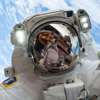 A NASA astronaut above with the Earth behind them and the ISS reflected in their helmet visor