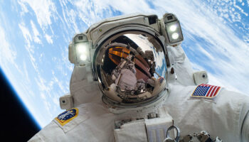 A NASA astronaut above with the Earth behind them and the ISS reflected in their helmet visor
