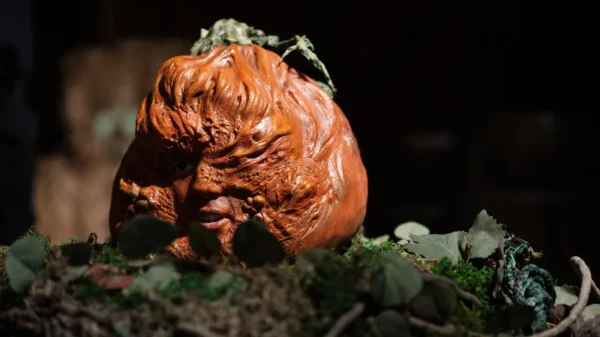 A deformed pumpkin head resembles a human face, it sits atop the soil