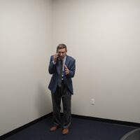 Tim Robinson is dressed in a suit and glasses, standing in the corner on a room with blank white walls, shouting into a mobile phone