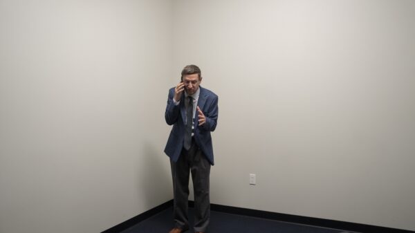 Tim Robinson is dressed in a suit and glasses, standing in the corner on a room with blank white walls, shouting into a mobile phone