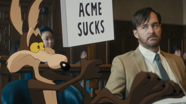 Cartoon Coyote in a real world court room with his human lawyer, holding up a sign which says "Acme Sucks", a still from Coyote vs. Acme